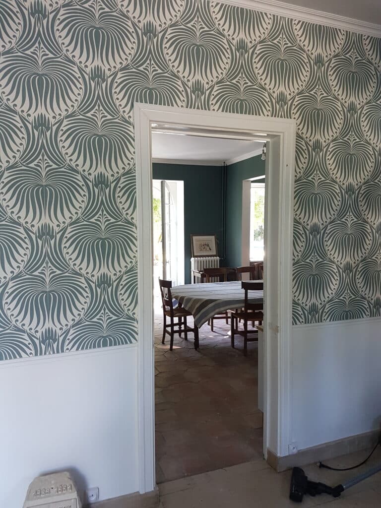 Papier peint Art Nouveau vert canard pour un intérieur classique. Papier peint Art Nouveau vert canard à motifs stylisés au-dessus d'une boiserie blanche, encadrant une porte vers une salle à manger aux murs sombres.