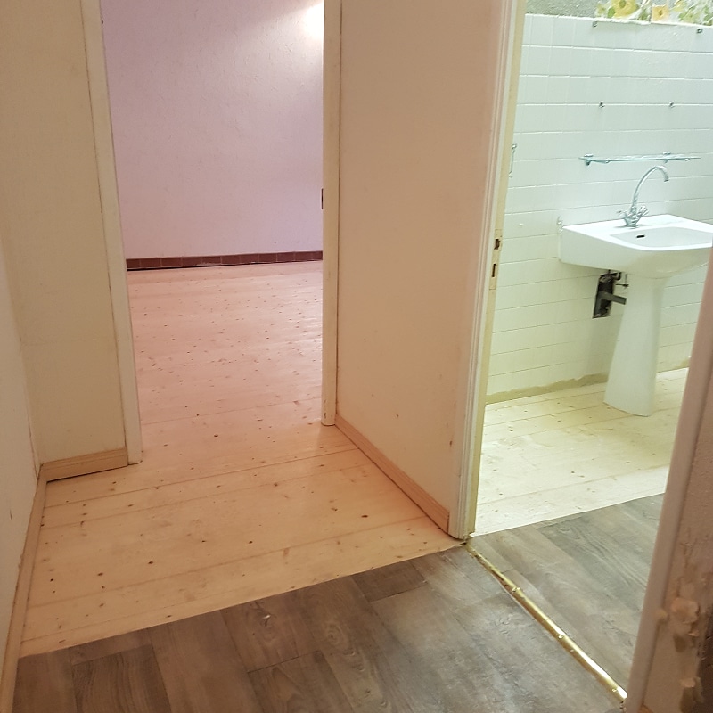 Rénovation intérieure : transition sol bois clair vers vinyle foncé. Transition entre sol vinyle et plancher bois clair brut dans un couloir. La vue mène à une chambre aux murs roses et à une salle de bain avec lavabo blanc.