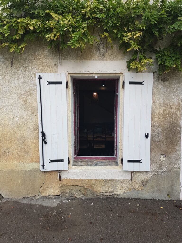 Charme rustique: Fenêtre avec volets blancs sur façade ancienne. Volets blancs ouverts sur une vieille façade en crépi beige sous une vigne verte. L'intérieur sombre est faiblement éclairé par une lampe suspendue.