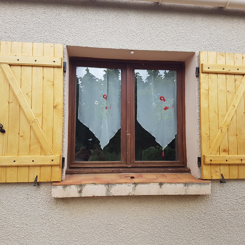 Fenêtre encadrée par des volets en pin clair à barre et écharpe ouverts. Rideaux blancs ornés de coquelicots sur une façade en crépi.